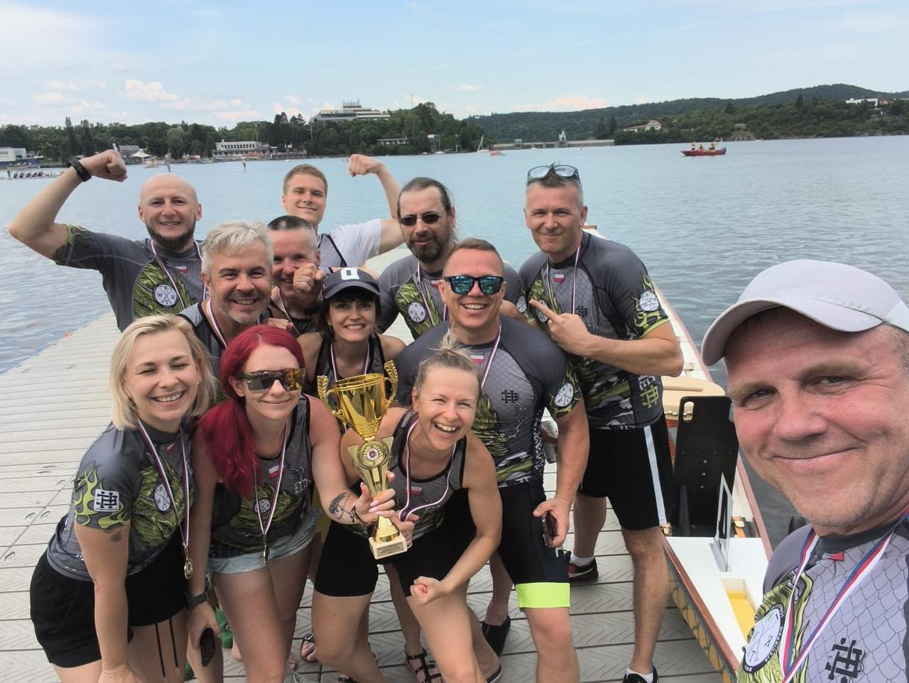 Uśmiechnięta drużyna KTW z medalami i złotym pucharem na pomoście nad zbiornikiem w Brnie, jeden z wioślarzy robi selfie, Festival dračích lodí Brno 2026