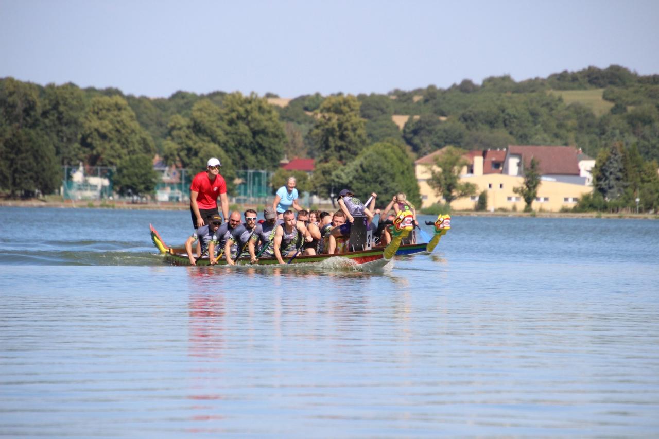 Smocza łódź KTW płynie po spokojnym jeziorze w słońcu, osada w szarych koszulkach w jednym rytmie, sternik w czerwonej koszulce stoi na rufie, Olšovec Open 2026