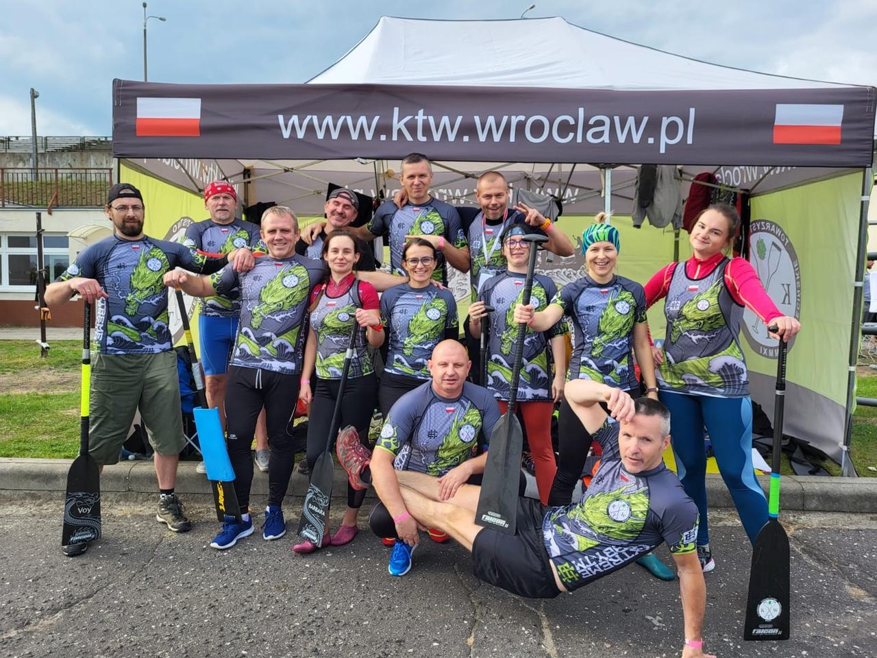 Drużyna KTW w klubowych strojach ze smokiem pod namiotem z adresem strony klubu, wioślarze z wiosłami